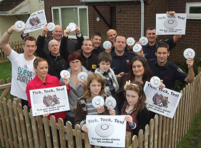 Friends, family, and firefighters pose with posters and smoke alarms in front of the house