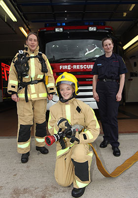 Two visitors and one firefighter pose in front of a fire appliance, the visitors are dressed in full protective kit