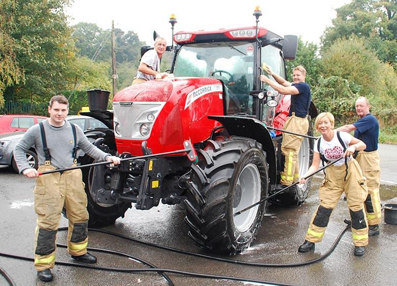 5 firefighters wash a big tractor on a tarmac drive with trees in the background